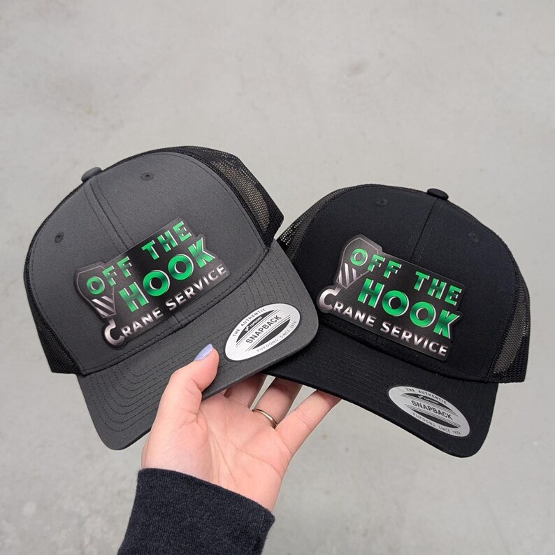 May include: Two snapback trucker hats, one dark gray and one black, featuring mesh back panels and a front patch with a crane hook graphic and the text "OFF THE HOOK CRANE SERVICE" in green and silver. Each hat has a round silver sticker on the brim.