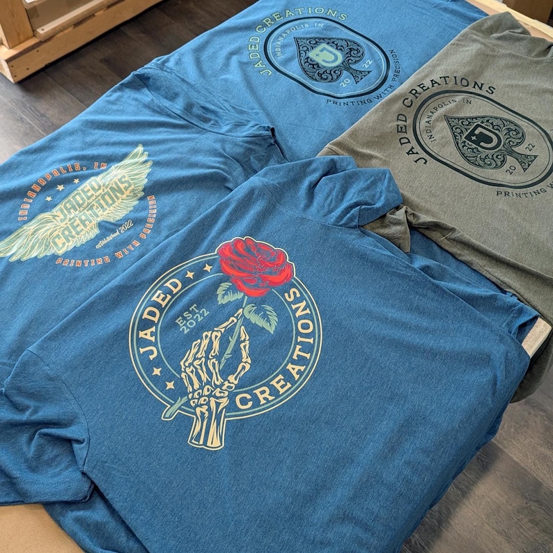 May include: A selection of blue and grey t-shirts, each with a unique graphic. One shirt has a red rose held by a skeleton hand, encircled by the text "JADED CREATIONS EST 2022." Other shirts feature a spade symbol and the words "JADED CREATIONS."