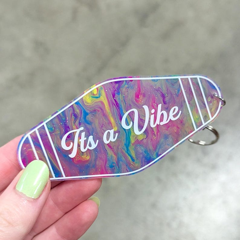 May include: A motel-style keychain with a colorful, swirling marble pattern in shades of pink, blue, and yellow. The keychain features white text that reads "Its a Vibe" in a cursive script, accented by white vertical stripes and a metal ring.