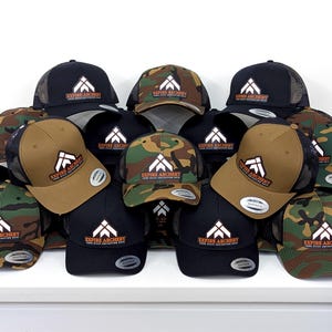 May include: A collection of snapback baseball caps in black, tan, and camouflage patterns. Each cap features a white geometric logo above the text "EXPIRE ARCHERY" and "YOUR HILLS ASPIRATION HATS" in orange. The hats have structured crowns and mesh backs.
