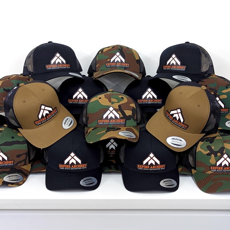May include: A collection of snapback baseball caps in black, tan, and camouflage patterns. Each cap features a white geometric logo above the text "EXPIRE ARCHERY" and "YOUR HILLS ASPIRATION HATS" in orange. The hats have structured crowns and mesh backs.