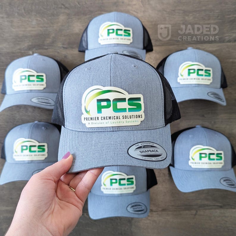 May include: A collection of grey and black trucker-style snapback caps featuring a white patch with the green and blue PCS Premier Chemical Solutions logo. Each cap has a structured front panel, a mesh back, and a flat brim with a sticker.