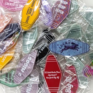 May include: A collection of assorted motel-style keychains in individual plastic sleeves, featuring various colors, patterns, and printed text including Poppy's Taxi Service, Lucky with a rabbit graphic, 3 Migos, and 132 Saint Marks Place Apartment 7.
