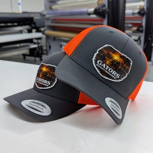 Op de afbeelding: Twee grijze en oranje baseballpetten met de tekst "GATORS RESOURCE MANAGEMENT" op een patch. De petten hebben een gebogen klep en een mesh rug. De petten zijn gemaakt door Snapback.