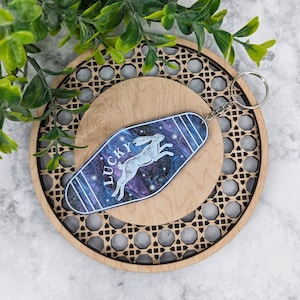 Op de afbeelding: Een sleutelhanger in motelstijl met een donkerblauwe en paarse gemarmerde achtergrond, een witte afbeelding van een springend konijn en het woord LUCKY in witte letters. De sleutelhanger heeft een zilverkleurige metalen ring aan de bovenkant.