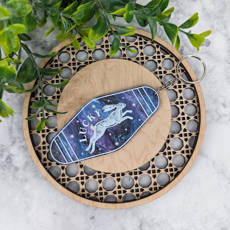 Op de afbeelding: Een sleutelhanger in motelstijl met een donkerblauwe en paarse gemarmerde achtergrond, een witte afbeelding van een springend konijn en het woord LUCKY in witte letters. De sleutelhanger heeft een zilverkleurige metalen ring aan de bovenkant.
