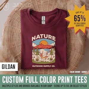 Könnte beinhalten: Ein granatrotes T-Shirt mit Rundhalsausschnitt und einem Grafikdruck, der einen Hirsch zeigt, der in einer Hängematte an einem See entspannt, mit Bergen und Sonne im Hintergrund. Der Text lautet NATURE OUTDOOR SUPPLY CO.