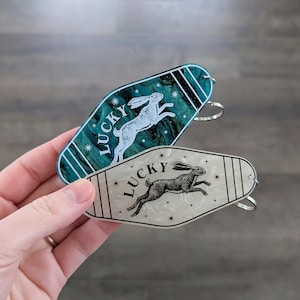 Op de afbeelding: Twee sleutelhangers in motelstijl met een puntige ovale vorm. De ene is donkerblauwgroen met een witte konijnillustratie en het woord LUCKY. De andere is cr&egrave;mekleurig met een zwarte konijnillustratie en het woord LUCKY. Beide hebben steraccenten.
