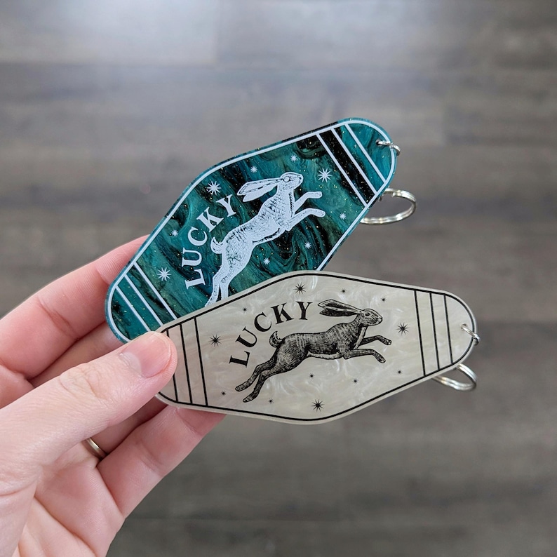Op de afbeelding: Twee sleutelhangers in motelstijl met een puntige ovale vorm. De ene is donkerblauwgroen met een witte konijnillustratie en het woord LUCKY. De andere is cr&egrave;mekleurig met een zwarte konijnillustratie en het woord LUCKY. Beide hebben steraccenten.