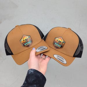 May include: Two tan snapback hats with black mesh back panels. Each hat features a shield-shaped patch on the front displaying a sunset landscape, silhouettes of three people, and the text POM ADVENTURES. A silver sticker is on the brim.