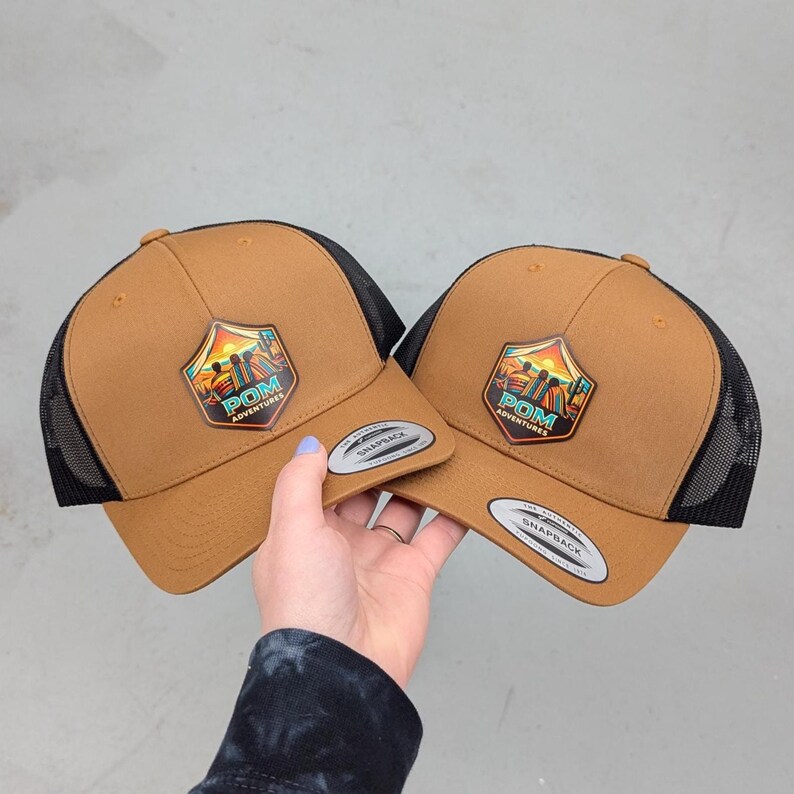 May include: Two tan snapback hats with black mesh back panels. Each hat features a shield-shaped patch on the front displaying a sunset landscape, silhouettes of three people, and the text POM ADVENTURES. A silver sticker is on the brim.