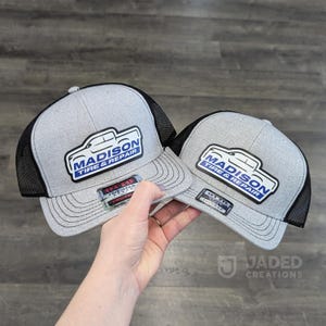 May include: Two grey and black trucker caps featuring a white patch with a blue and black graphic of a pickup truck and the text Madison Tire & Repair. Each cap has a flat brim with stitching and a small rectangular sticker on the front.