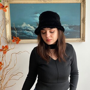 May include: A woman wearing a black long-sleeved shirt, blue jeans, and a black faux fur bucket hat. The hat has a wide brim and a soft, fuzzy texture.