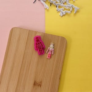 May include: Two pink cookie cutters on a wooden cutting board. One cutter is shaped like a nutcracker and the other is shaped like a princess.