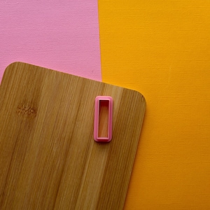 May include: A wooden cutting board with a pink rectangle-shaped cookie cutter on top. The cutting board is on a pink and orange background.