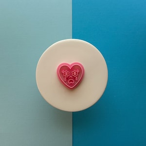 May include: Pink heart-shaped cookie cutter with a crying face and flower eyes. The cutter is on a white surface.
