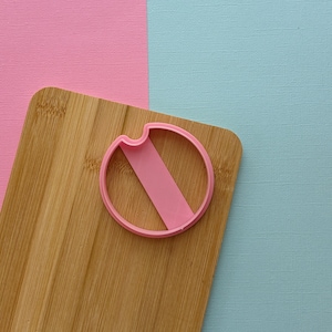 May include: A pink cookie cutter in the shape of a circle with a horizontal line across the center, sitting on a wooden cutting board.