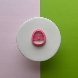 May include: Pink plastic cookie cutter shaped like a small purse or handbag. The cutter has a small handle on top and a detailed design.