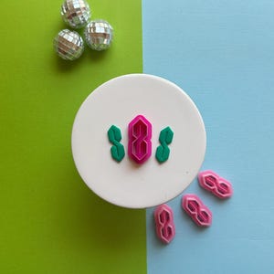 May include: A set of pink and green plastic cookie cutters shaped like the number 8, with a unique, stylized design. The cutters are on a white plate, with three more pink cutters on a blue surface.