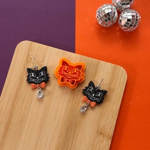 May include: Black cat earrings with orange bow ties and dangling crystal accents are displayed on a wooden cutting board. An orange cat-shaped cookie cutter and three disco balls are also present. The background is split between purple and orange.