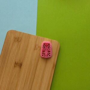 May include: A pink plastic cookie cutter with a decorative design on a wooden cutting board.