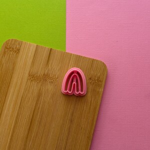 May include: A pink plastic cookie cutter shaped like an archway, sitting on a wooden cutting board.
