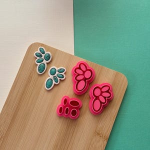 May include: A collection of colorful, decorative embellishments. Two are turquoise with white borders, and three are shades of pink. They are arranged on a wooden cutting board against a white and teal background.