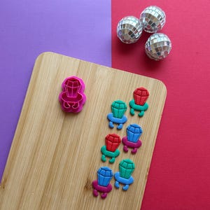 May include: A pink plastic cookie cutter shaped like a ring with a diamond on top. There are eight colorful rings made from fondant on a wooden cutting board. The rings are red, blue, green, and turquoise.