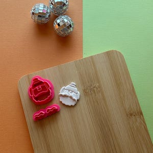 May include: Three silver disco balls and three cookie cutters in the shape of Santa Claus and a mustache are arranged on a wooden cutting board. The background is divided into orange and green sections.