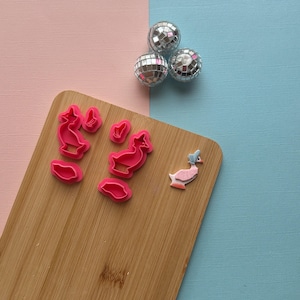 May include: A set of pink plastic cookie cutters shaped like ducks, with a pink duck cookie on a wooden cutting board.
