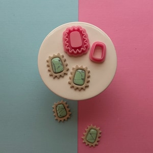May include: Four green and brown resin cabochons with a marbled pattern, and two pink plastic molds for making jewelry on a white plate.
