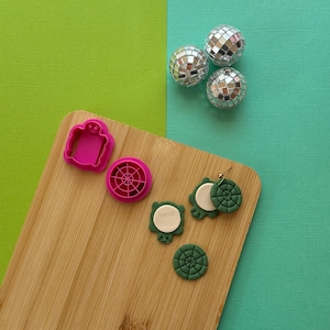 May include: A wooden cutting board with two pink cookie cutters and four green clay earrings. The cookie cutters are shaped like a turtle and a circle with a design. The earrings are also shaped like turtles and circles with a design. There are three silver disco balls in the background.