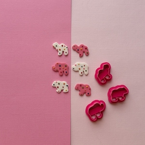 May include: Pink and white cookie cutters in the shapes of bears and camels. The cutters are on a pink and white background.