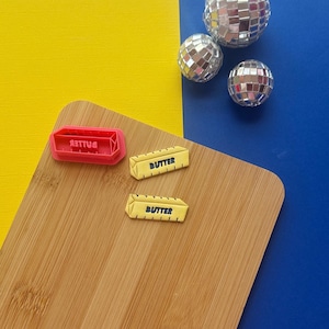 May include: A wooden cutting board with a pink rectangular mold that says "BUTTER" in reverse, and two yellow butter-shaped objects with the word "BUTTER" printed on them. Three disco balls are in the background.