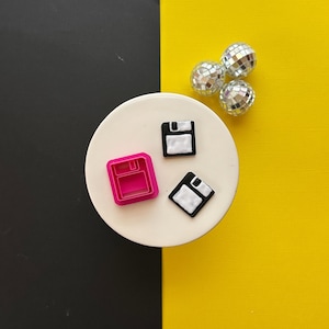 May include: Three floppy disk shaped cookie cutters, one pink, two black and white, on a white surface.