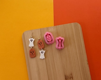 May include: Two pairs of Halloween-themed earrings on a wooden surface. One pair features a white ghost charm and a brown pumpkin charm. The other pair features a white ghost charm and a brown pumpkin charm. There are also two pink Halloween-themed cookie cutters, one shaped like a ghost and the other shaped like a pumpkin.