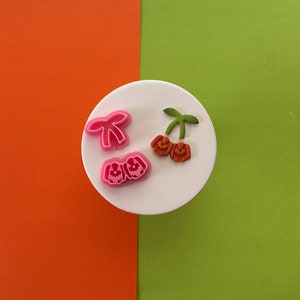 May include: A set of three pink and orange plastic cookie cutters. The cutters are shaped like a bow, two shoes, and a cherry.