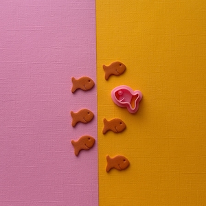 May include: A pink fish-shaped cookie cutter stands out against a yellow background, surrounded by five orange fish-shaped cookie cutters on a pink background.