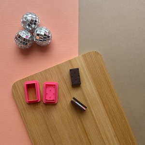 May include: A wooden cutting board with two pink cookie cutters, one chocolate and one pink and white cookie, and three silver disco ball ornaments on a pink and brown background.