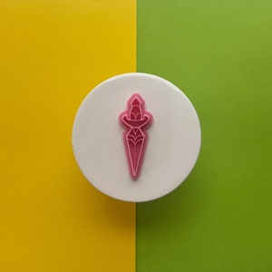 May include: A pink plastic dagger cookie cutter on a white background.