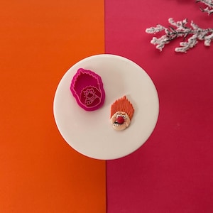 May include: Two pink silicone cookie cutters, one shaped like the Grinch and the other shaped like the Grinch's hair. The cutters are on a white plate.