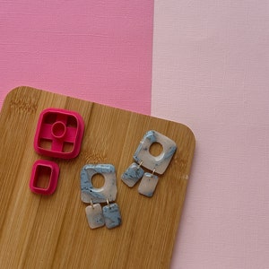 May include: Two pairs of gray and pink marbled square earrings with a hole in the center. The earrings are on a wooden surface with two pink plastic square cookie cutters.