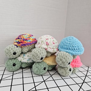 May include: A collection of handmade crochet turtle plushies. Each turtle features a textured, rounded body and head, with small black eyes. The shells are in various colors and patterns, including floral, rainbow, and solid blue.