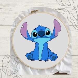 May include: A cross stitch pattern featuring Stitch from Disney's Lilo & Stitch. The pattern shows Stitch sitting with his signature blue fur, big eyes, and purple ears. The pattern is framed in a wooden embroidery hoop.