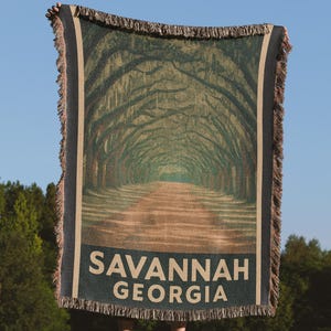 Puede incluir: Una manta tejida con una imagen de una avenida bordeada de árboles, con el texto "SAVANNAH GEORGIA" en la parte inferior. La manta tiene un borde con flecos y se sostiene contra un cielo azul y árboles verdes.