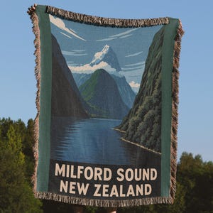 May include: A woven blanket depicting Milford Sound, New Zealand, with mountains and water. The text "MILFORD SOUND NEW ZEALAND" is displayed in white. The blanket has a green border and fringed edges. The scene is set against a blue sky.