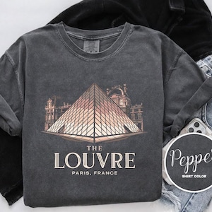 May include: Dark grey sweatshirt with a graphic of the Louvre Pyramid in Paris, France. The text "THE LOUVRE" and "PARIS, FRANCE" are printed below the image. The sweatshirt is styled with black trousers and blue jeans.