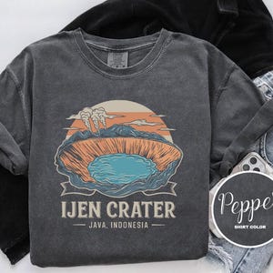 Puede incluir: Una sudadera gris oscuro con un gráfico del cráter Ijen en Java, Indonesia. El diseño presenta un lago azul, paredes de cráter naranjas y marrones y una puesta de sol. El texto "IJEN CRATER" está debajo.