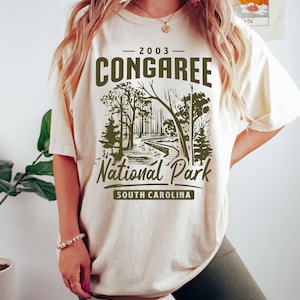 May include: A beige t-shirt with a green graphic design that says "2003 Congaree National Park South Carolina". The graphic features a stylized image of a forest path.