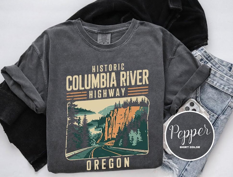 A gray t-shirt with a graphic design depicting the Historic Columbia River Highway in Oregon, along with a denim jacket and a pair of jeans.